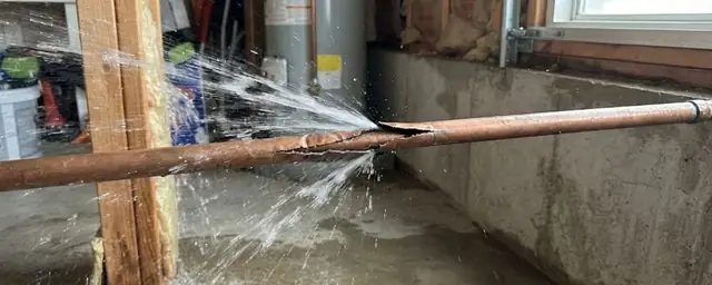 Split copper pipe spraying water across a basement floor